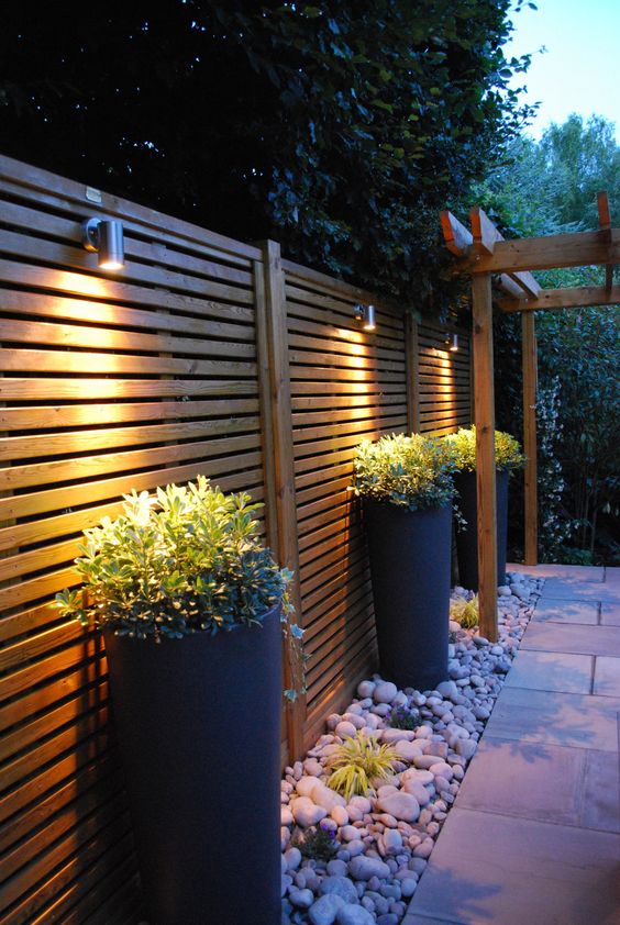 a stained fence with tall planters with greenery and wall lamps over them and rocks on the ground are a super chic and modern combo