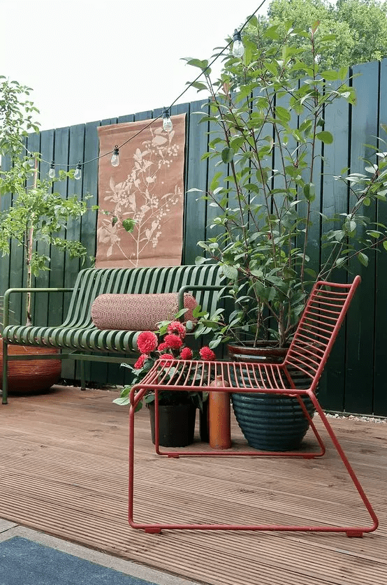 a stylish and bright terrace with a tall fence in green, a green metal sofa and a red chair, some potted plants and greenery