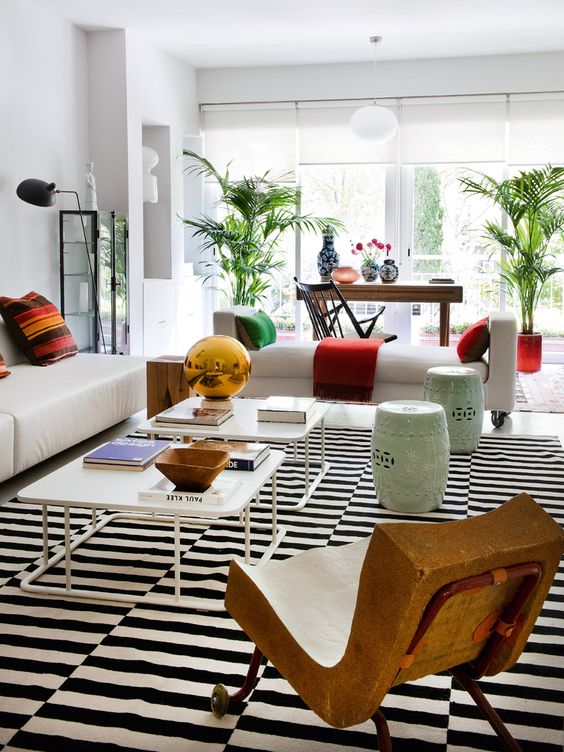 a stylish living room with a stripe rug, a white sofa and daybed, coffee tables, stools, potted plants, a desk with vases