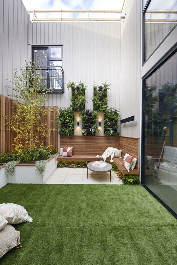 a stylish modern garden with a green lawn, a terrace with a built-in bench, some wall planters, trees and grasses