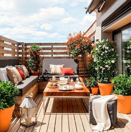 a summer balcony with a wooden deck and furniture, potted blooms and greenery and bright pillows plus candle lanterns