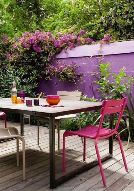 a terrace with a purple wall, a stained table, grey and pink chairs, some blooming vines is amazing