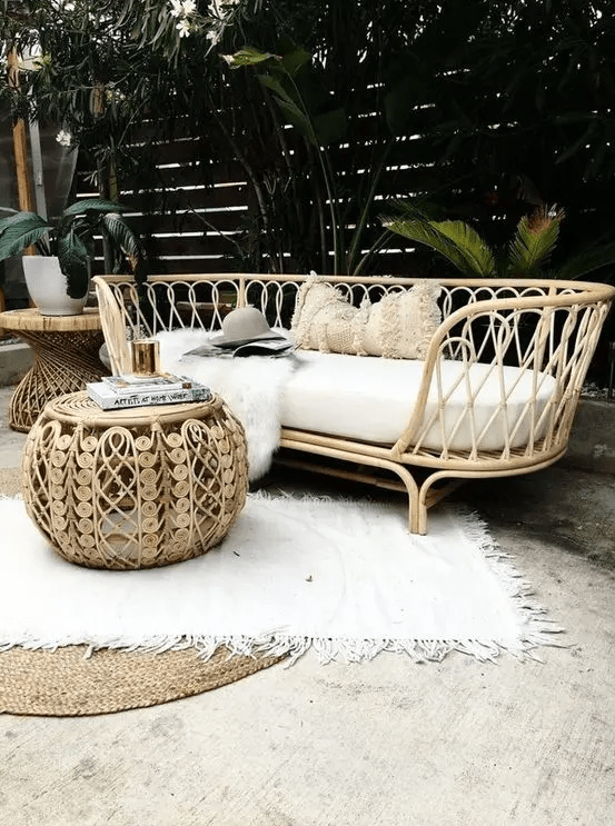 a tropical outdoor space with a rattan daybed, a rattan pouf and a side table, layered rugs and tropical plants around