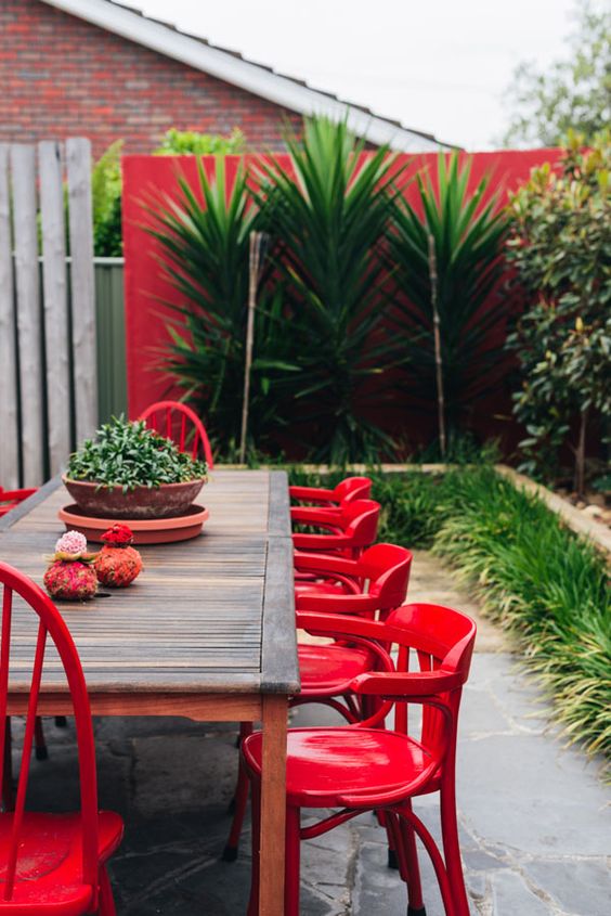 a tropical outdoor space with a stained table, red chairs, greenery and tropical plants along the red wall
