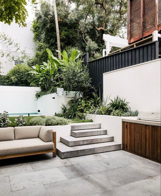 a two-level outdoor space with a pool and greenery, a sofa and an outdoor kitchen is a lush tropical space