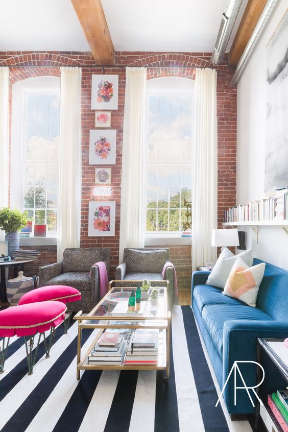 a whimsical living room with red brick walls, a blue sofa, a coffee table, pink stools and printed chairs, a striped rug and a gallery wall