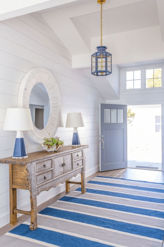 an amazing coastal entryway with white planked walls, a blue and grey striped rug, a wooden console table and blue touches