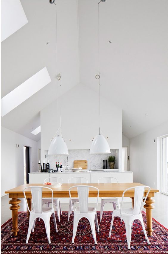 an eat-in kitchen with white cabinets, pendant lamps, a bold boho rug, a stained table and white chairs plus some skylights