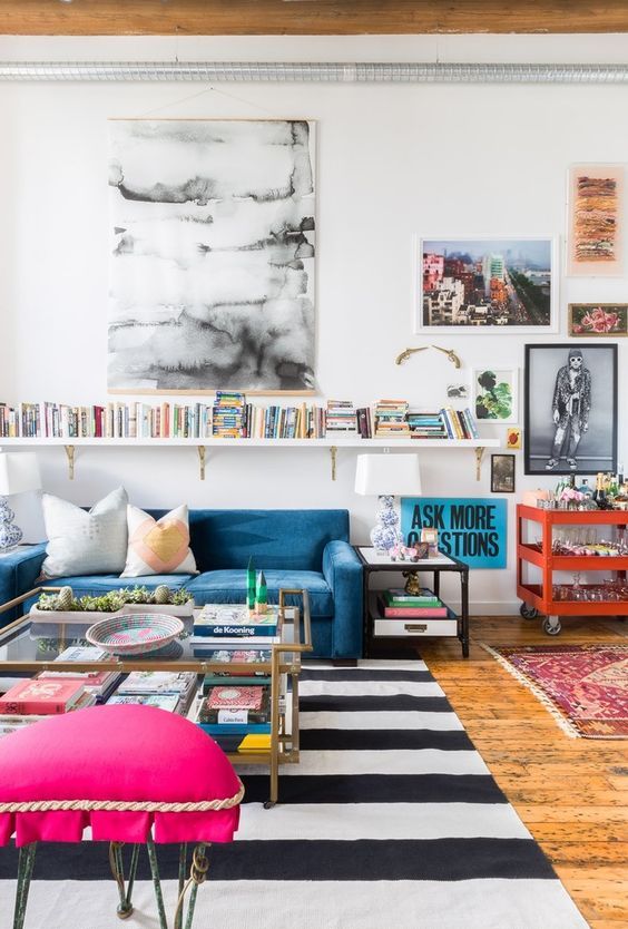 an eclectic living room with a blue sofa, a striped rug, a coffee table with books and a pink stool, a bookshelf and some artwork