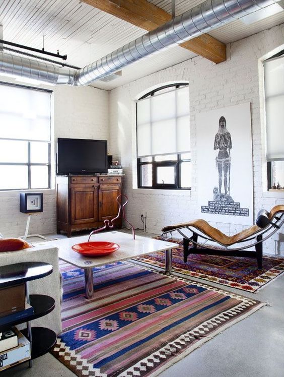 an eclectic living room with birck walls and exposed pipes, bright boho rugs, a daybed, a coffee table and a white sofa