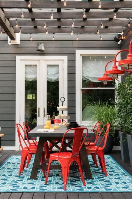 an outdoor dining space with a table, red metal chairs, a bodl blue rug and red wall lamps and some potted greenery