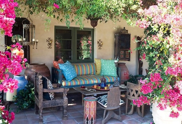 A Moroccan boho terrace with plenty of color, with dark stained carved furniture, colorful pillows and upholstery, greenery and bold flowers