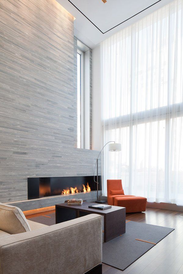 A double height minimalist living room with a wood clad fireplace wall, contemporary furniture and a large window for more natural light