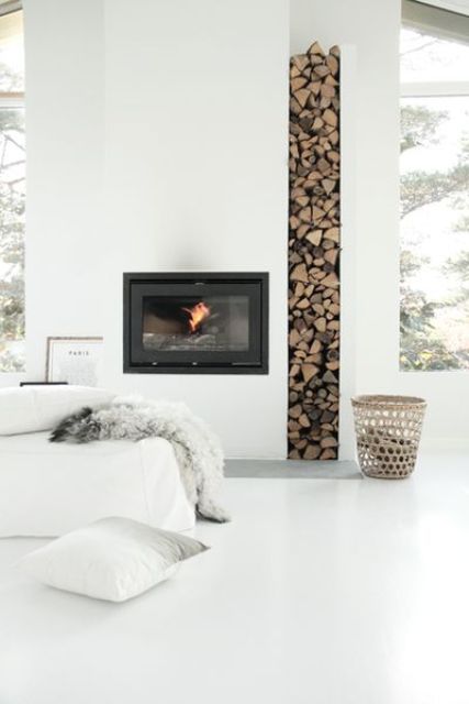 A minimalist all white living room with a fireplace and a firewood storage space, a white sofa and large windows for more light