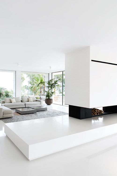 A minimalist and light filled living room with a fireplace and firewood storage, a grey sectional sofa and panoramic windows