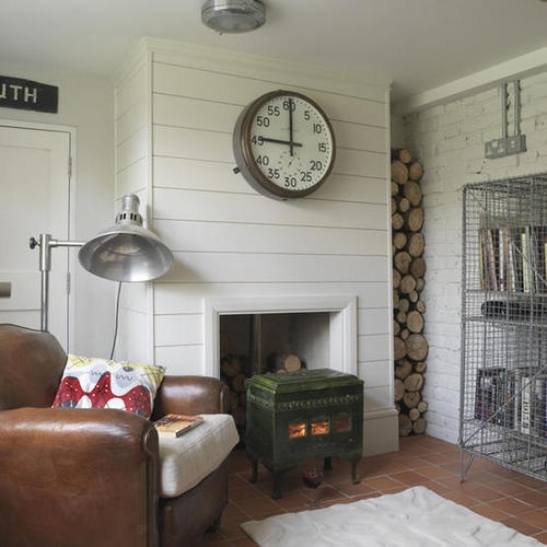 a neutral rustic living room with shiplap on the walls, a hearth and firewood plus some retro furniture