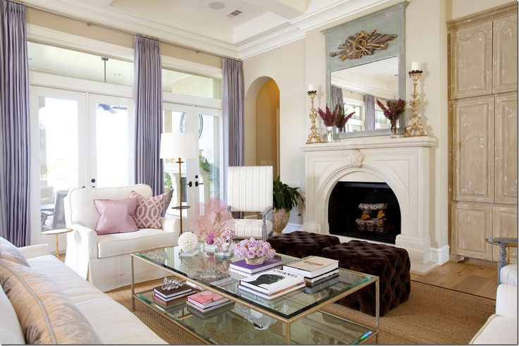 A glam vintage inspired neutral living room with a fireplace, white seating furniture, lilac curtains, touches of pink and a glass coffee table