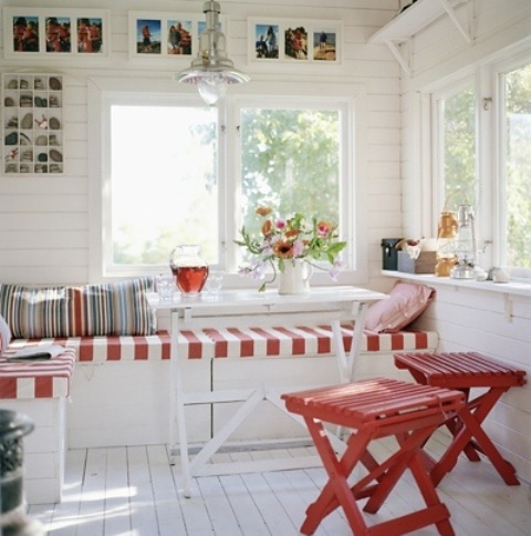 A white Scandinavian sunroom with a built in bench with striped upholstery, red stools and a dining table and a gallery wall