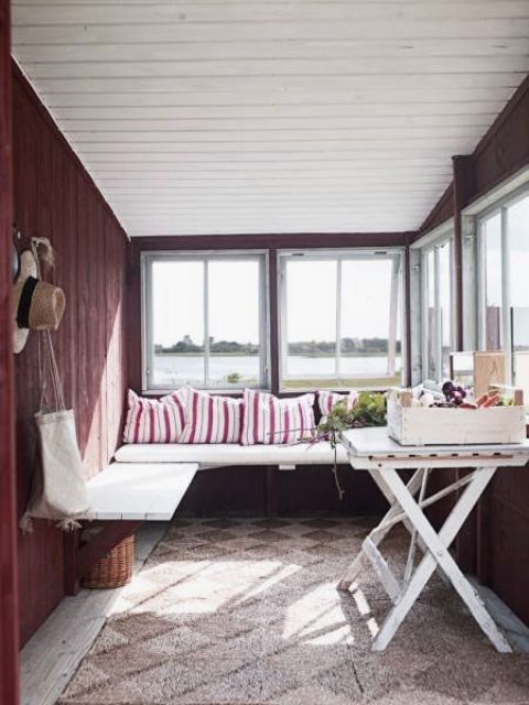 A burgundy Scandinavian sunroom with a built in bench, a white trestle table and some striped pillows and some accessories