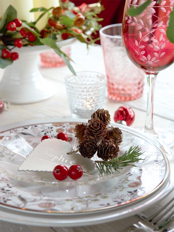 An elegant Christmas place setting with printed porcelain, red glasses and berries, pillar candles and colored candleholders