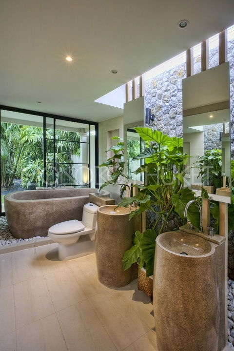 A lush tropical bathroom with stone and glass walls, a stone tub, free standing sinks and lots of statement tropical plants
