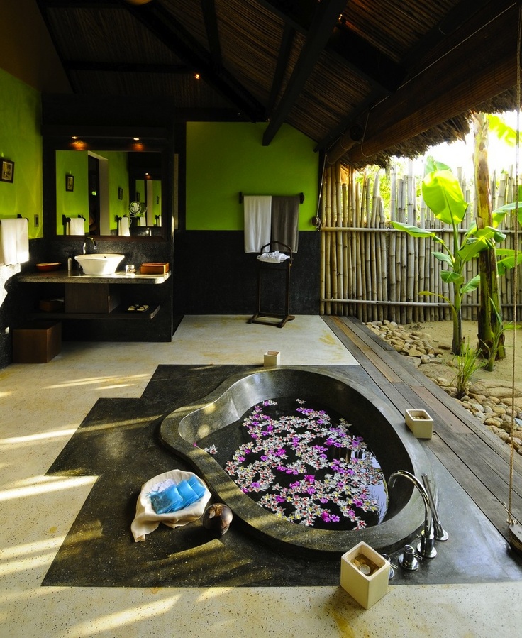A bright indoor outdoor bathroom with green and black walls, a sunken stone tub with a view to outdoors and a glass wall that can be removed