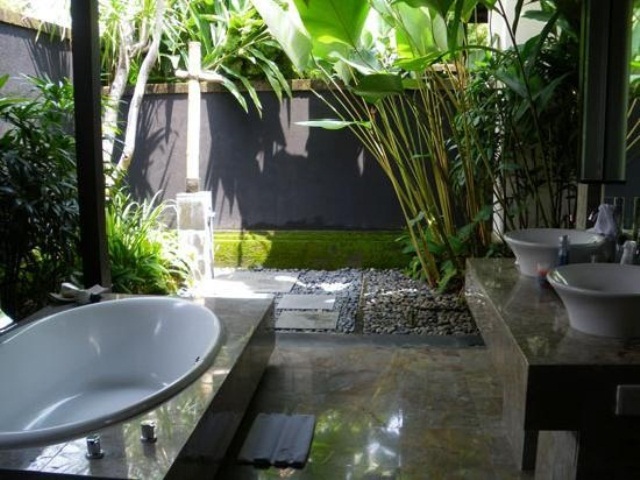 An outdoor indoor tropical bathroom with pebbles and growing tropical plants and a shower outside, a bathtub inside and sinks on a stone vanity