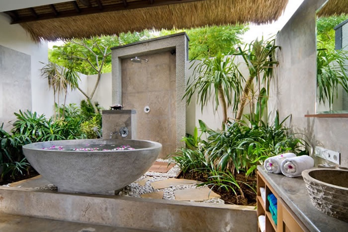 An outdoor indoor tropical bathroom clad with neutral stone, with a stone tub and tropical plants growing around plus stone sinks
