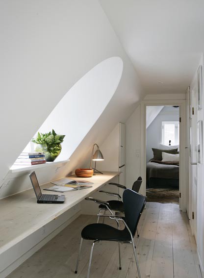 A small white attic home office with an arched window, a long built in floating desk, a built in storage cabinet and some artworks on the wall