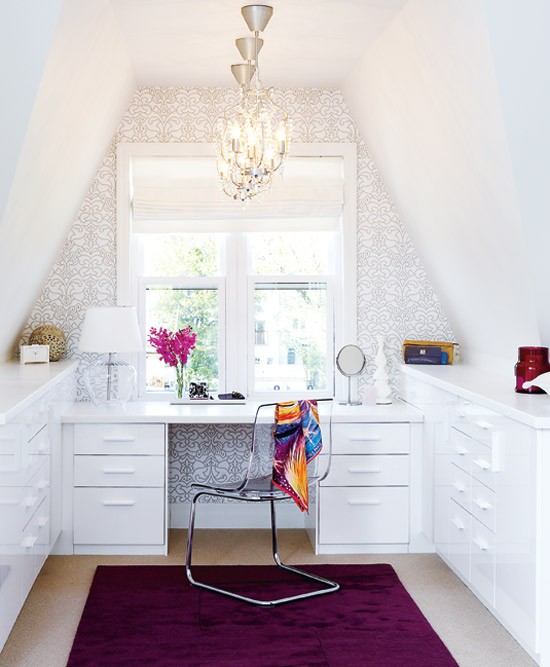 A glam attic home office with two storage units and a built in desk, an accent wall done with printed wallpaper, chic glam chandeliers, an accent purple rug and a sheer chair
