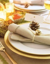 a place setting with a gold charger, a white plate, a neutral napkin with a pinecone and a bell is very chic and Christmassy