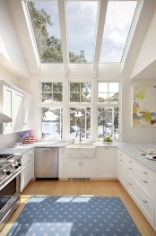 a large white kitchen with shaker cabinets, a large window and a skylight that allow to enjoy a lake view and some trees for relaxing