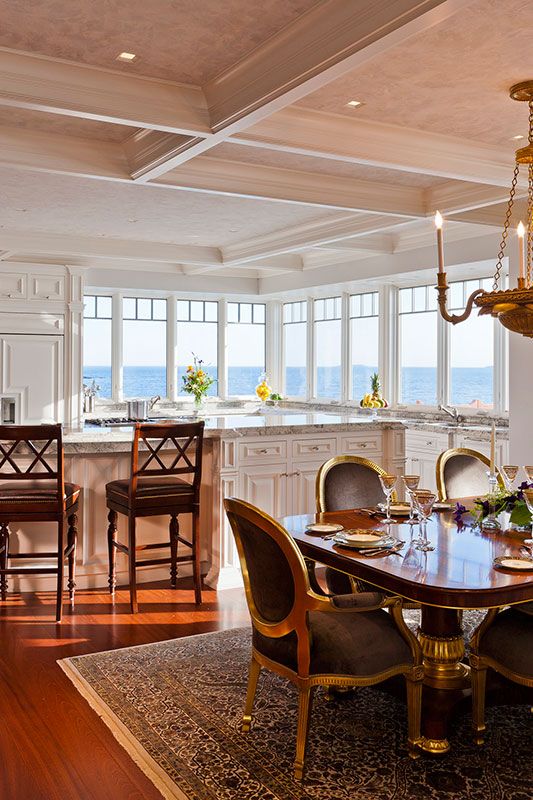 a vintage white farmhouse kitchen with shaker cabinets, white stone countertops, large windows with a sea view and a refined vintage dining space