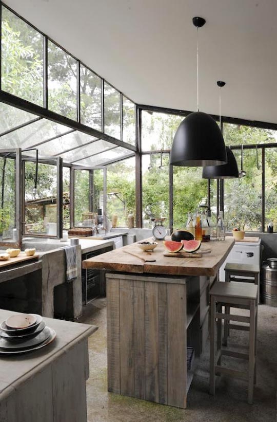 A natural indoor outdoor kitchen with glazed walls, pendant lamps, rough wooden furniture and lots of concrete