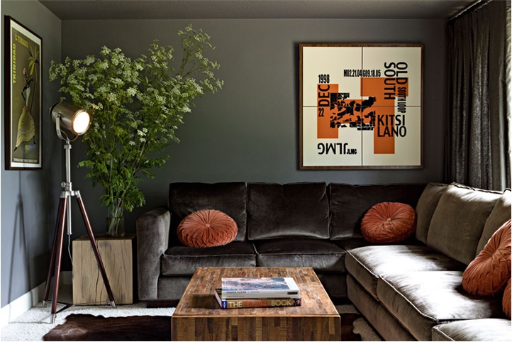A stylish dark living room with dark walls, an L shaped sofa, orange pillows and a graphic artwork, a floor lamp and a wooden coffee table