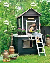a stylish black and white kids’ house with a raised deck, a ladder, greenery and blooms around is a nice example of Scandi style