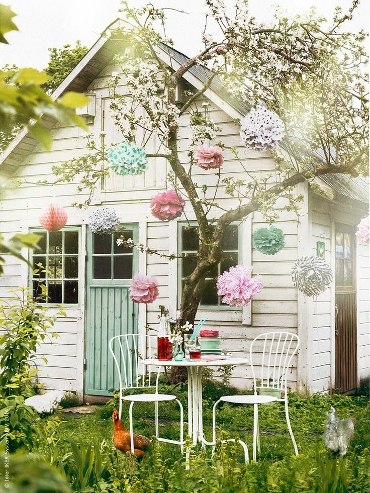 A white kids' playhouse with window frames and an aqua door, a tree with pastel pompoms and a metal dining table and chairs around
