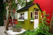 a bright green mini kids’ playhouse with a black roof, white window frames and a door, with greenery and a small pond