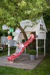 a raised white kids’ playhouse with a ladder and a red slide, with potted blooms and greenery is a great spot for kids’ games