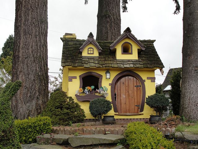A tiny fairytale like kids' playhouse in yellow, with a dark roof, dark window frames and a cute door reminds of hobbits' houses