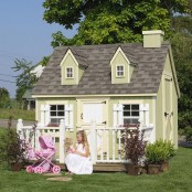 a small green kids’ playhouse with a dark roof, white window frames and a door, potted plants and a pink baby carriage