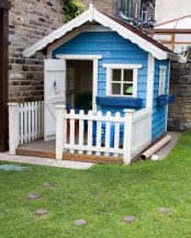 a blue and white kids’ playhouse with a white door and window frames, a small porch with a fence is cute and bright