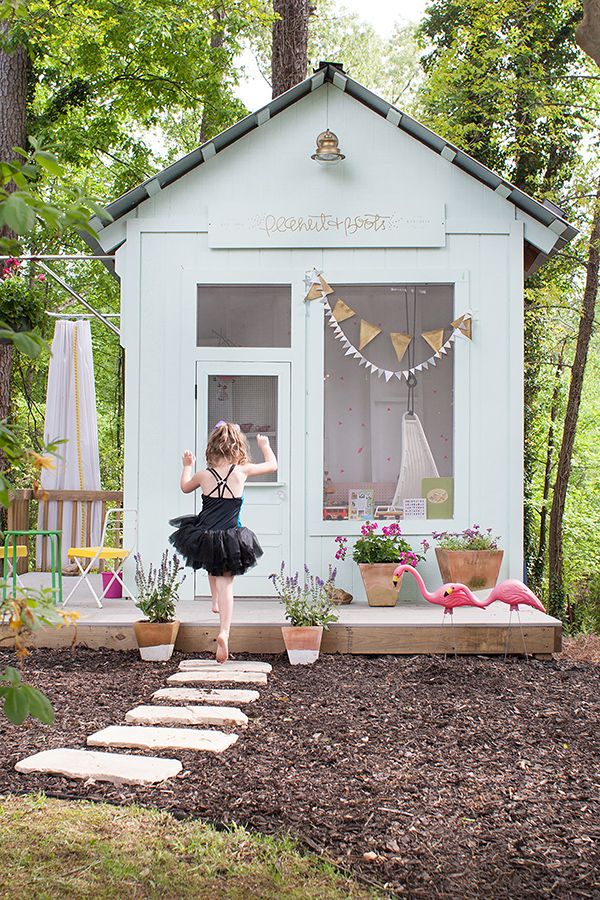 A whitewashed kid's playhouse with a large window, some cool furniture inside, outdoor furniture and potted blooms plus a flamingo