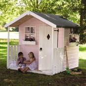 a light pink kids’ playhouse with a white door and windows, with a small porch and potted blooms and gardening tools around