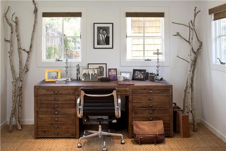 A neutral rustic home office with a wooden desk, tree branches, a jute rug, burlap shutters and a stylish mid century modern chair