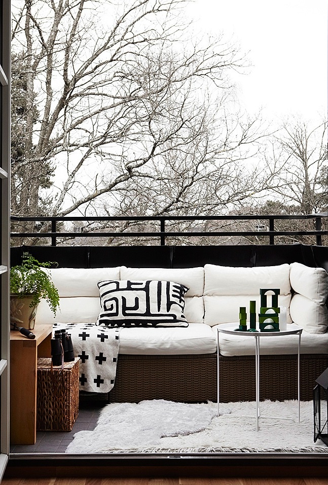 A Nordic balcony with a wicker sofa with white upholstery, a wood and wicker side table, greenery and a glass side table