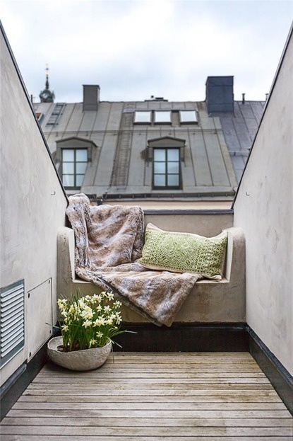 A very small Scandinavian balcony with a built in seat with pillows and a blanket, potted blooms is a simple and chic space to stay