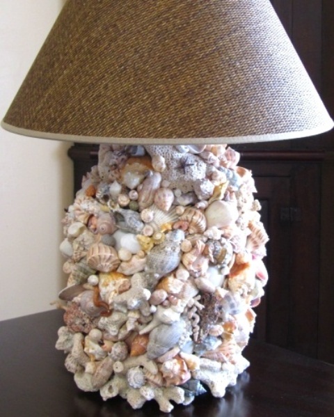A sea inspired table lamp with a base covered with seashells of various colors and sizes and a burlap lampshade for a rustic seaside home