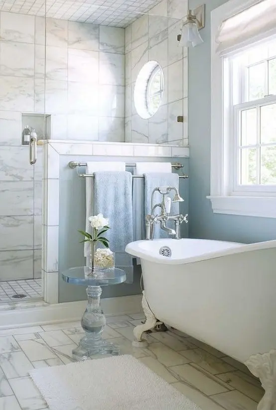 a beautiful bathroom with blue walls and a shower space clad with marble tiles, with a vintage bathtub and a pony wall in the shower space
