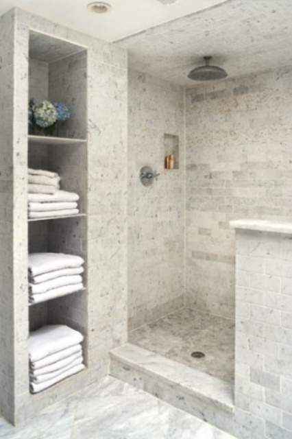 A neutral contemporary bathroom clad with white marble tiles, with a half wall for the shower space and built in shelves